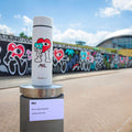 A limited edition Nuby RapidCool x Mul white thermal flask with cartoon hearts and characters stands outdoors, matching a graffiti mural. A sign highlights its RapidCool technology. Only 500 units made.
