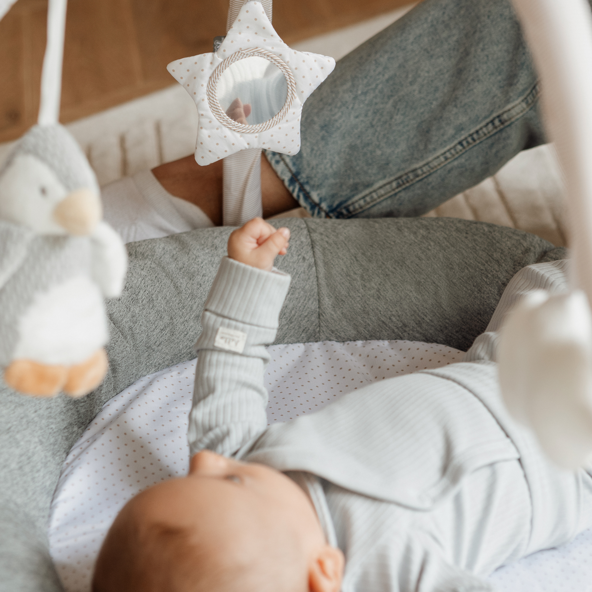 A baby plays on the Nuby Penguin and Pals Play Gym, reaching for a star-shaped mirror toy, while a plush penguin hangs nearby and an adult’s legs are seen in the background.