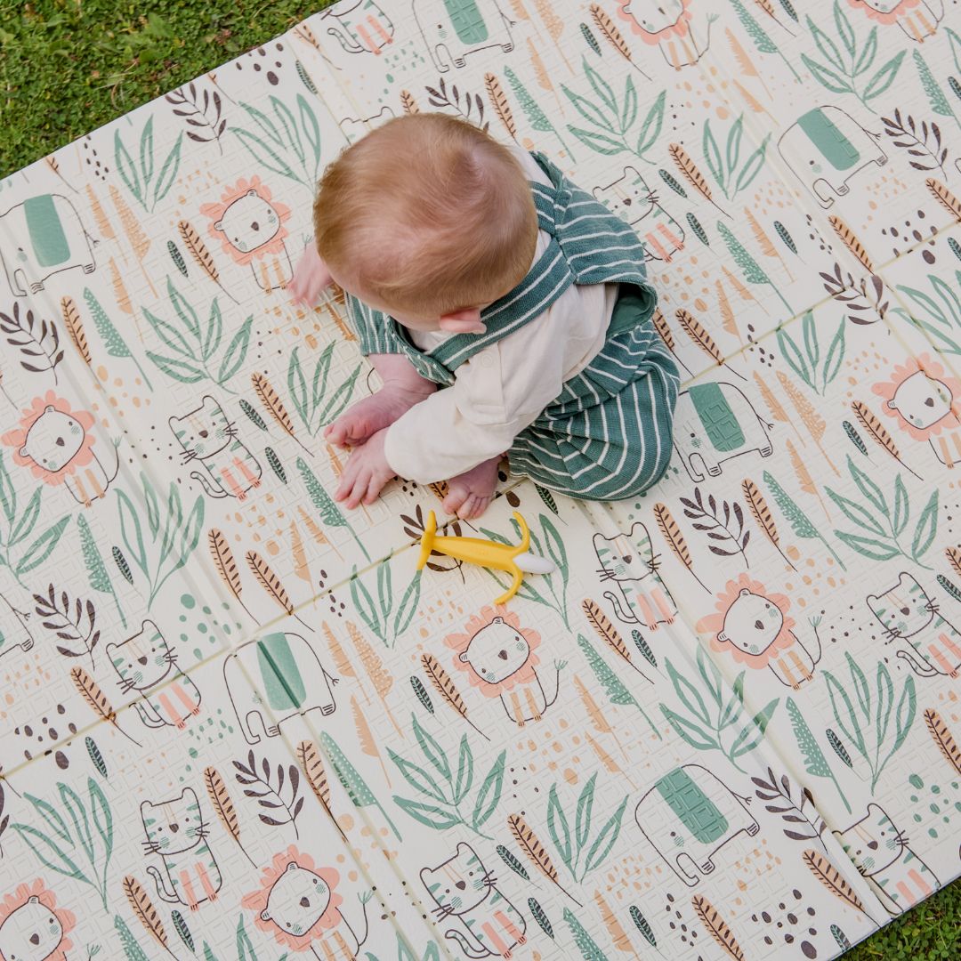 A baby in green striped overalls sits on the Fold N Go Play Mat, which features animal illustrations and leafy patterns, reaching for a yellow toy. The foldable play mat offers a soft, cushioned surface for play, with grass visible in the top left corner.