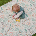 A baby in green striped overalls sits on the Fold N Go Play Mat, which features animal illustrations and leafy patterns, reaching for a yellow toy. The foldable play mat offers a soft, cushioned surface for play, with grass visible in the top left corner.
