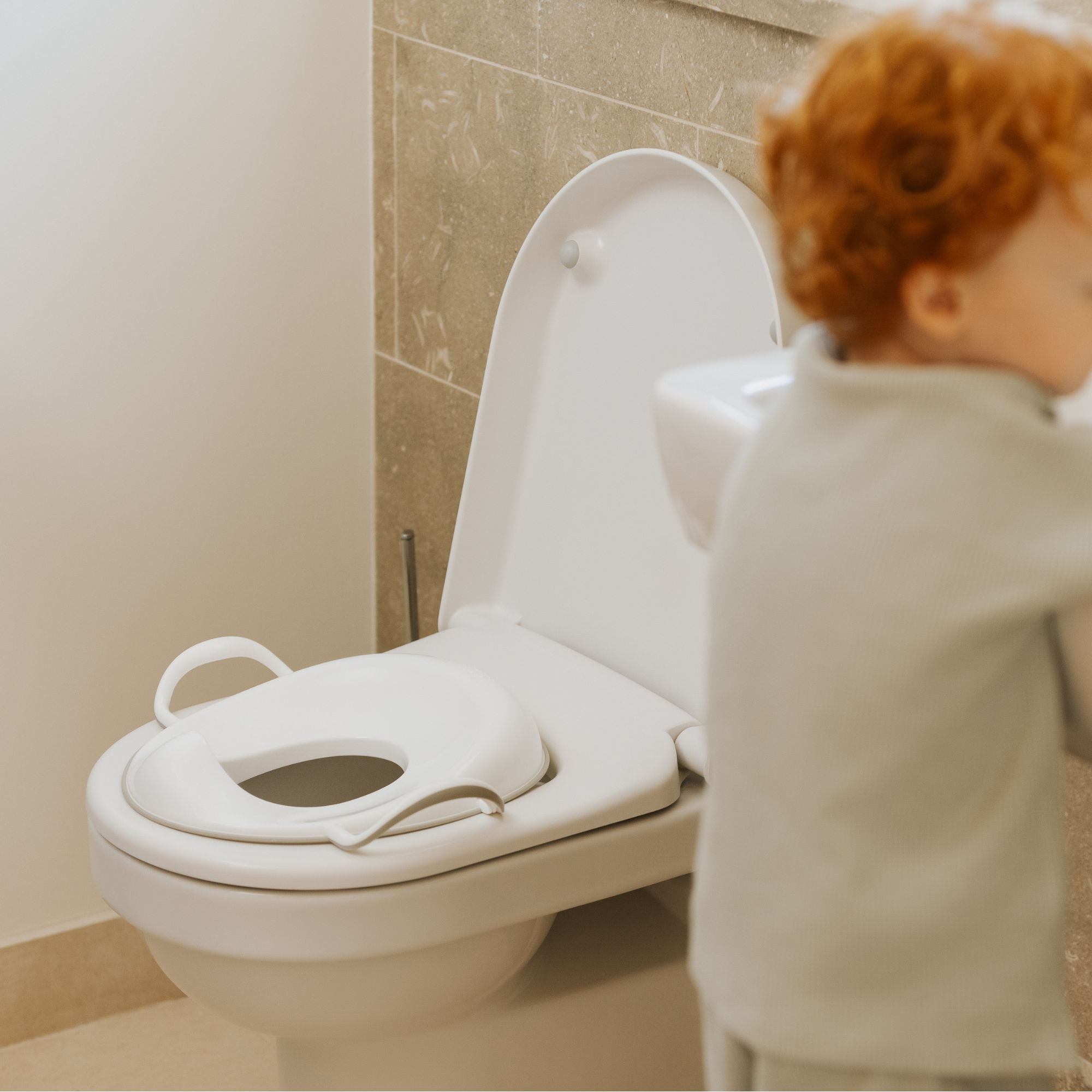 A Potty Training Kit Bundle by Nuby UK is attached to a white toilet. A young child with curly red hair stands nearby, facing away. The background has beige tiled walls.