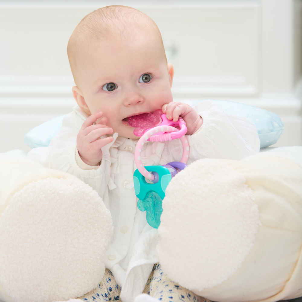 A baby in a white outfit sits supported by pillows, holding and chewing on the Icy Bite Keys Teether Toy, looking up with wide eyes.