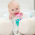 A baby in a white outfit sits supported by pillows, holding and chewing on the Icy Bite Keys Teether Toy, looking up with wide eyes.