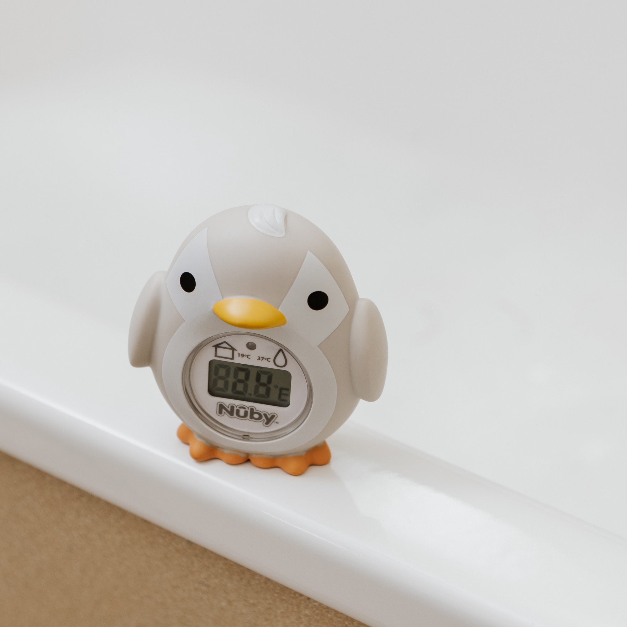 The Nuby UK Newborn Bath Support & Thermometer is a gray, penguin-shaped baby bath device with orange feet and beak. Its LCD screen displays water temperature, making it perfect for monitoring bath time for newborns.