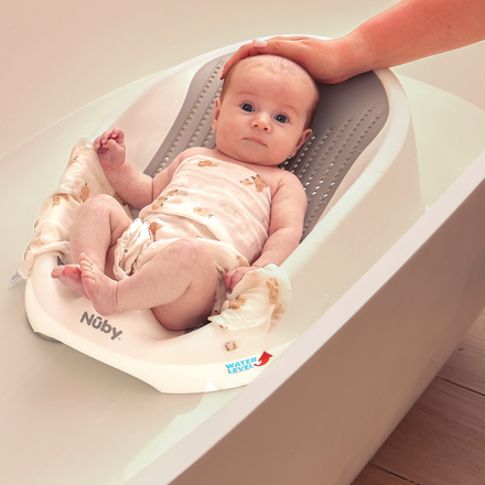 A baby rests comfortably in the Newborn Baby Bath Support, which features a cushioned backrest and is placed inside a bathtub filled with water. An adult hand gently cradles the baby’s head as they gaze upward in a light patterned onesie. The Newborn Baby Bath Support ensures safety and comfort during bath time, while its maximum water level indicator helps maintain the ideal bathing depth.