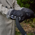 A person wearing Hand Pram Mittens, which are black and fur-lined, along with a beige coat, holds the pushchair handle of a stroller outdoors amidst greenery and fallen leaves.