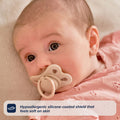 A baby in a pink outfit lies on a soft surface, using a beige RapidSoother dummy from the 2-pack of Dummies. A label reads: Hypoallergenic silicone-coated shield that feels soft on skin.