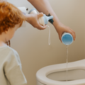 An adult pours liquid from a blue Nuby Gotta Go Travel Urinal into a toilet as a red-haired child watches, showcasing potty training with this leak-proof portable urinal in the bathroom.