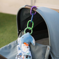 A close-up of a blue baby stroller shows a Nuby Linkables plush elephant toy hanging from the handlebar by colorful plastic links, dangling above the seat with grass visible in the background.