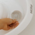 A hand holds a translucent, round item under water in a white container labeled Nuby Newborn Baby Bath, ideal for compact baby bathing.