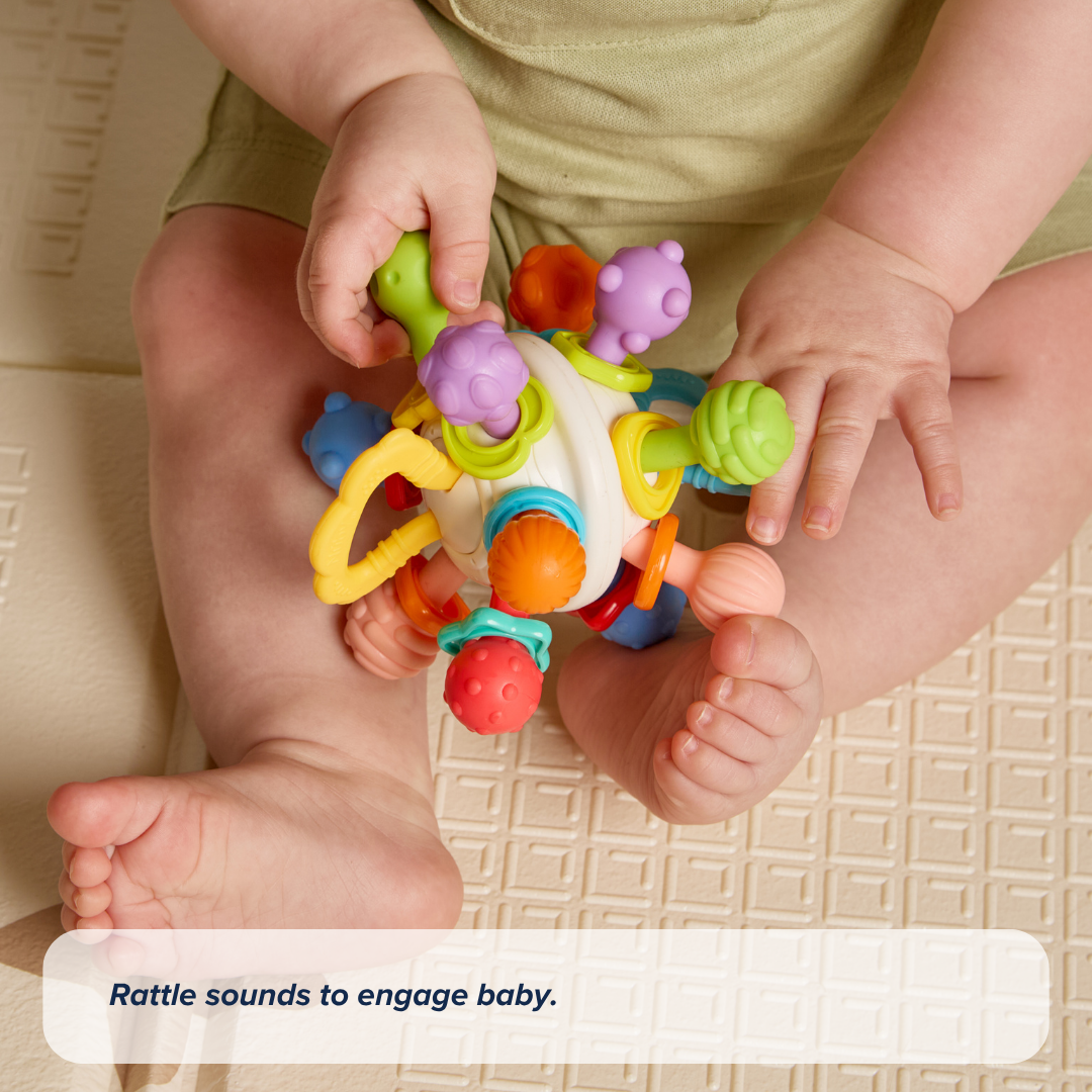 A baby sits on a mat, holding the Twist Ball Teether with both hands, as text below reads: Rattle sounds to engage baby.