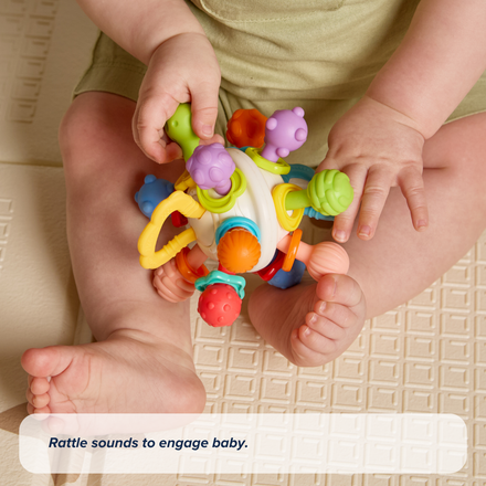 A baby sits on a mat, holding the Twist Ball Teether with both hands, as text below reads: Rattle sounds to engage baby.