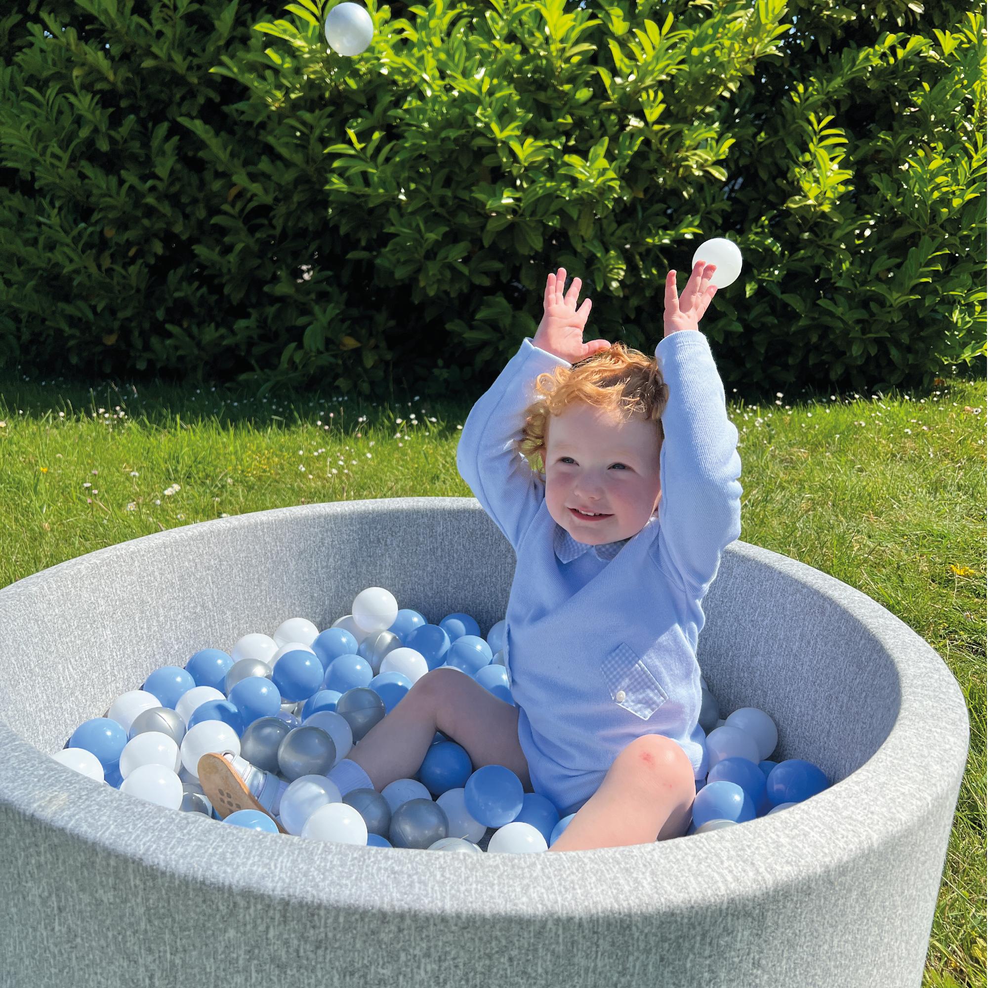 A smiling toddler in a blue outfit sits in the Ball Pit Play Set outdoors, tossing white balls into the air. The round ball pit, containing blue, white, and gray balls, is part of a soft play set with green bushes visible in the background.