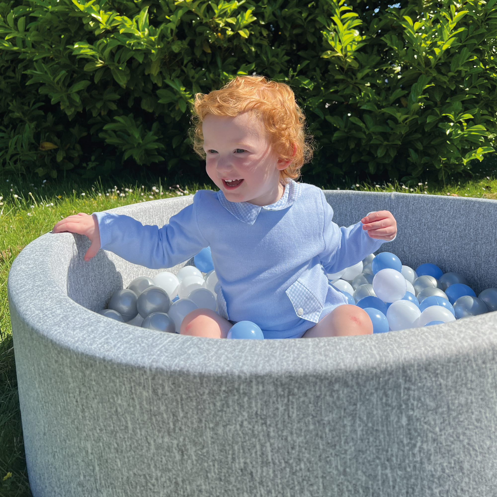 A smiling toddler with curly red hair enjoys the Ball Pit Play Set—a soft, safe gray ball pit filled with white, blue, and gray balls—while sitting outdoors on a sunny day.