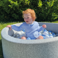 A smiling toddler with curly red hair enjoys the Ball Pit Play Set—a soft, safe gray ball pit filled with white, blue, and gray balls—while sitting outdoors on a sunny day.