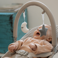 A baby in a beige outfit relaxes in the Nuby Musical Moves Baby Bouncer Chair, smiling at a plush moon and star mobile overhead. A blurred turquoise object is visible in the background.