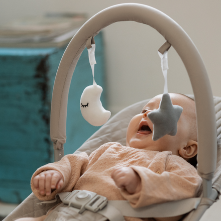 A baby in a beige outfit relaxes in the Nuby Musical Moves Baby Bouncer Chair, smiling at a plush moon and star mobile overhead. A blurred turquoise object is visible in the background.