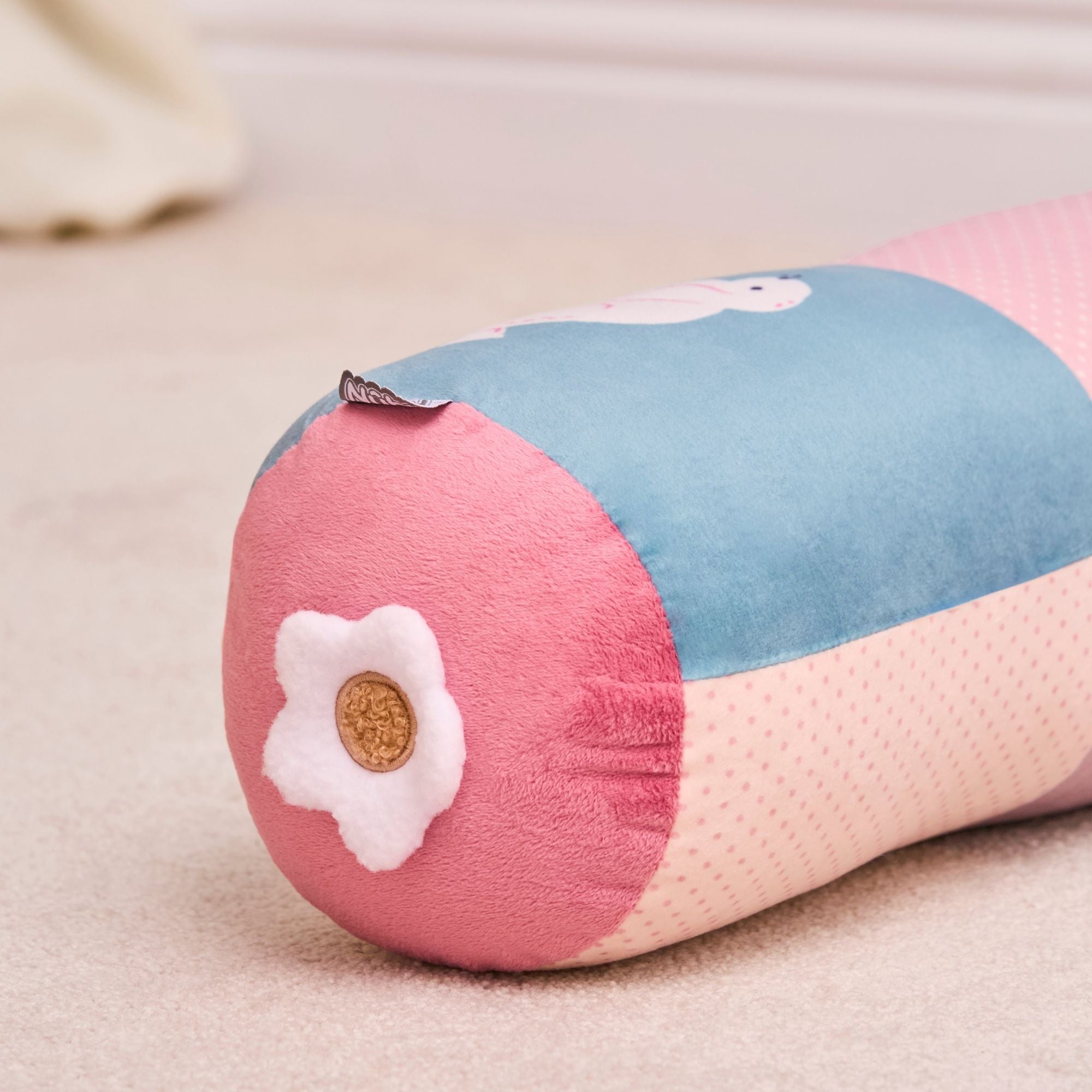 A close-up of the Little Hops & Raindrops Tummy Pillow, a cylindrical plush with pink, blue, and cream sections, featuring a white flower-shaped patch with a brown center on one end, resting on a beige carpet.