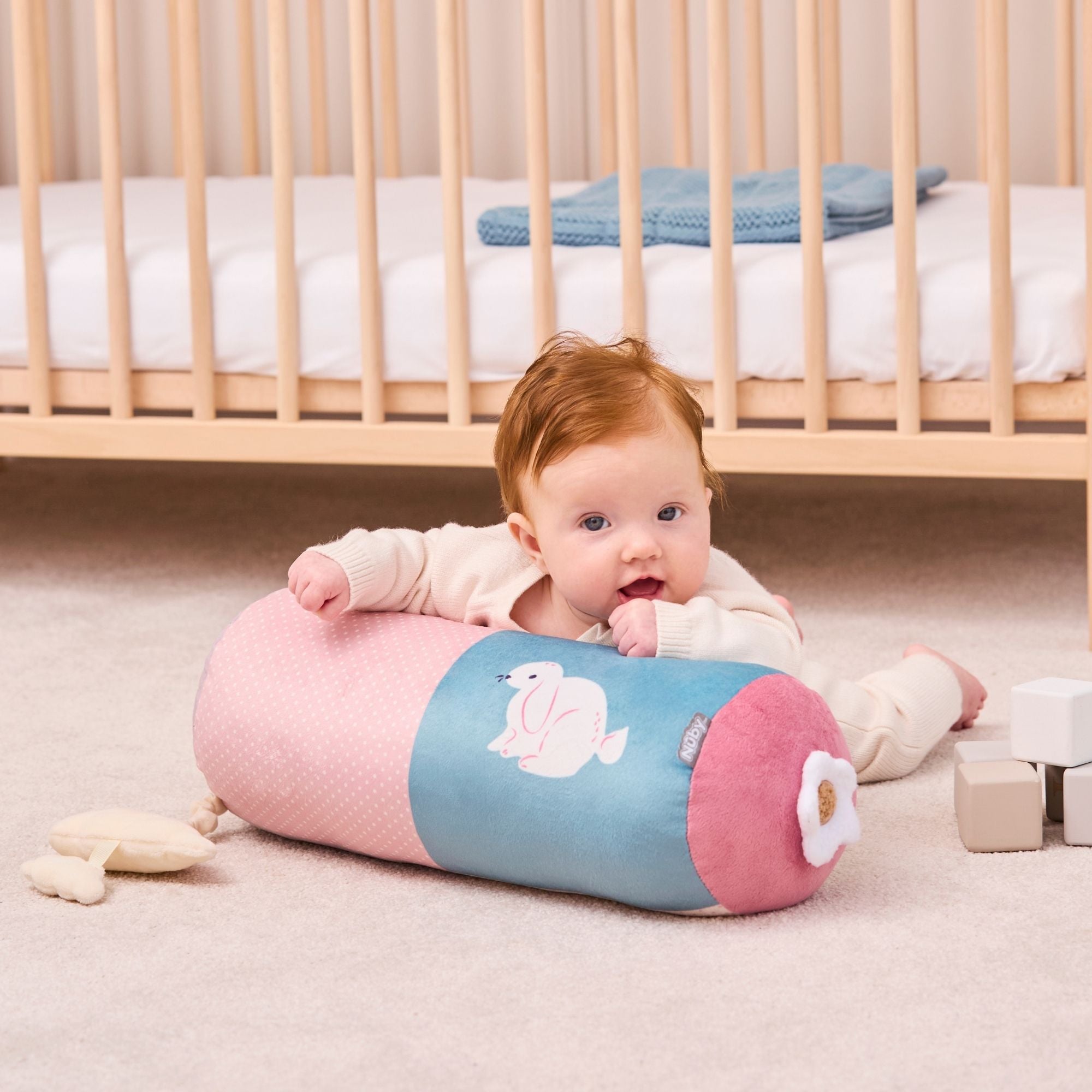 A baby with red hair lies on a soft carpet, propped up on the Little Hops & Raindrops Tummy Pillow near a wooden crib, soft toys, and pastel blocks.