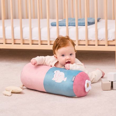 A baby with red hair lies on a soft carpet, propped up on the Little Hops & Raindrops Tummy Pillow near a wooden crib, soft toys, and pastel blocks.