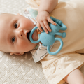 A baby in a pale yellow outfit lies on a patterned mat, holding and chewing the Nuby Blue Elephant Yogi Teether, featuring loops and handles.