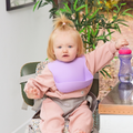 A blonde toddler in a topknot sits in a high chair wearing a pink outfit and one of the Silicone Bibs 3 Pack, reaching for a pink and purple sippy cup on a wooden table with plants in the background.
