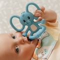 A baby lies down, gazing up with wide eyes while bringing the Nuby Blue Elephant Yogi Teether to their mouth. Wearing a patterned bib, they enjoy the comforting silicone teether.