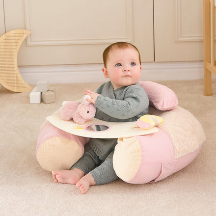 A baby in a grey outfit sits on a beige carpet, supported by the Little Hops & Raindrops Sit Up Seat with play tray. Holding a pink toy, the baby is surrounded by sensory toys and cozy décor in the room while looking up.