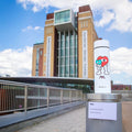 A limited edition Nuby RapidCool x Mul white water bottle featuring a colorful cartoon heart stands outdoors near the Baltic Centre for Contemporary Art, with a small sign in front of it.
