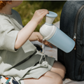 A child in a light green outfit sits outdoors, holding the Nuby Gotta Go Travel Urinal with a blue body, white top, and strap beside a gray backpack—ideal for families needing a leak-proof portable solution on any adventure.