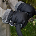 A person wearing a tan coat uses Hand Pram Mittens in black and gray, attached to a pushchair handle outdoors with grass and a wooden fence in the background.