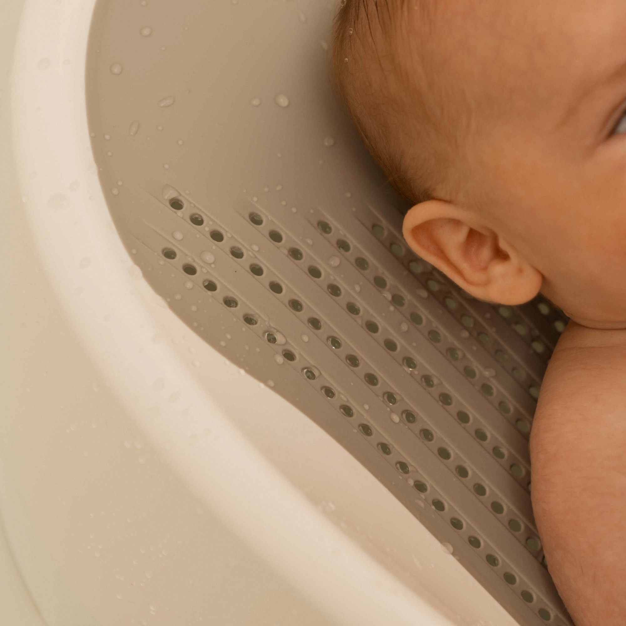 A close-up of a baby in the Nuby UK Newborn Bath Support & Thermometer, a beige tub with a textured non-slip surface and drainage holes. Water droplets cover the support, gently cradling the babys head, ear, and arm.