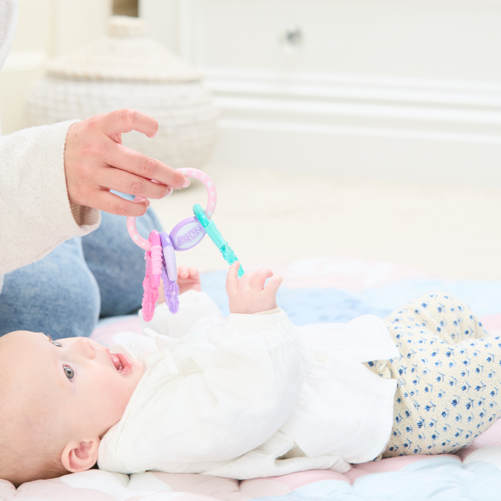 A baby dressed in a white shirt and patterned pants lies on a soft, pastel blanket, reaching excitedly toward the Icy Bite Keys Teether Toy held by an adult.