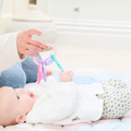 A baby dressed in a white shirt and patterned pants lies on a soft, pastel blanket, reaching excitedly toward the Icy Bite Keys Teether Toy held by an adult.