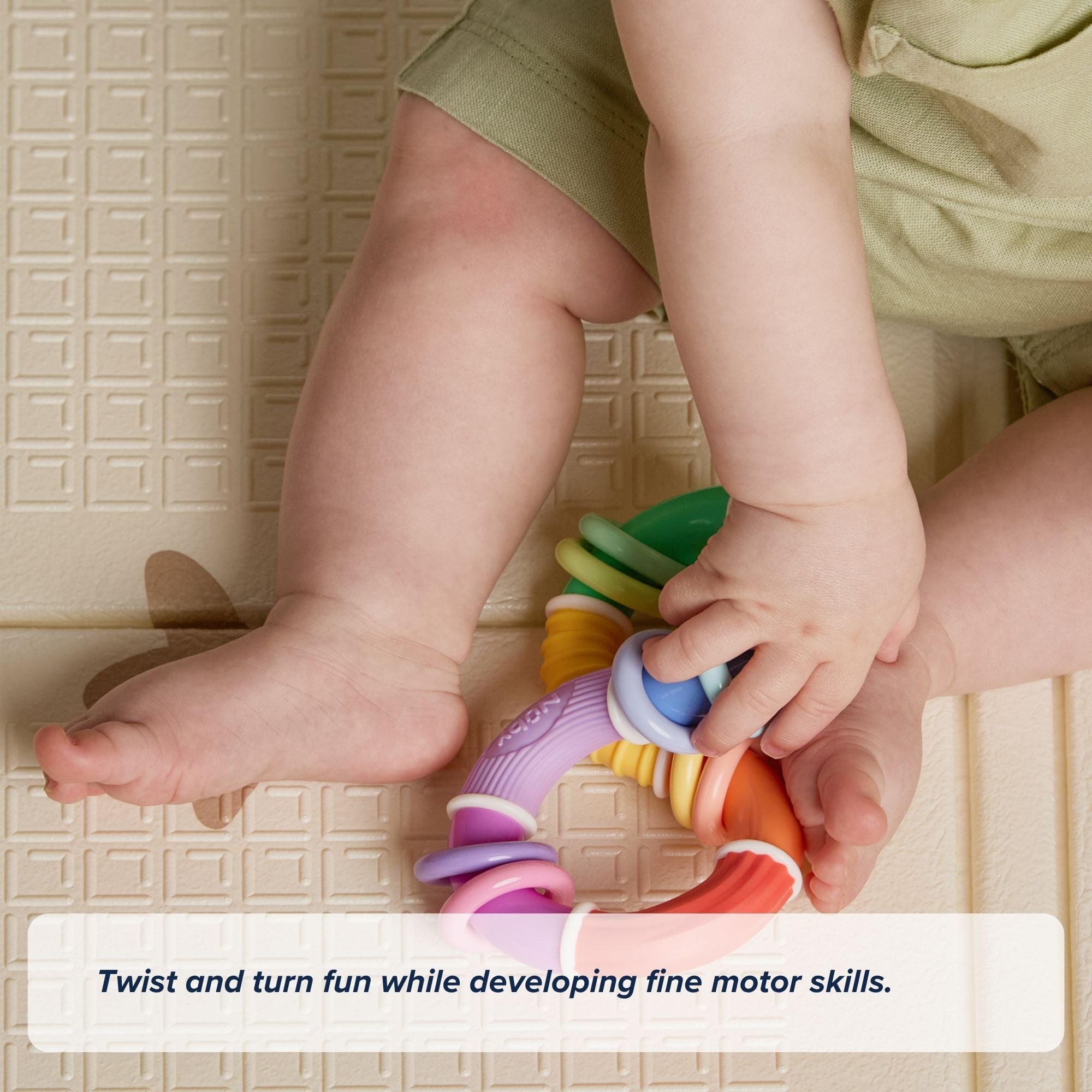 A baby sitting on a play mat holds and twists the Twista Rattle Teether with both hands, promoting fine motor skill development and offering teething relief.