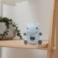 The Nuby Hippo Bath & Room Thermometer, shaped like a blue hippo and displaying 24°C, rests on a wooden shelf with a potted plant in the background.