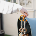 A hand holds two orange Nuby Linkables stroller hooks attached to a blue canopy, with baby toy rings and a soft ball hanging below. The person wears a white and gray striped sweater.