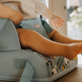 A small child sits in the Nuby Travel Booster Seat - Panda, showing its blue color with animal patterns. Their legs and feet are visible as they rest their hands on the seat’s sides, set against a softly blurred background.
