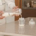 A person pours milk from a large Nuby bottle into a smaller Nuby baby bottle on the kitchen counter, with a Nuby breast milk storage bag and a bottle nipple lid nearby.