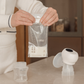 A person holds a Nuby Breast Milk Storage Bag filled with milk near a countertop displaying a Nuby electric breast pump and empty bottle, with a kitchen background and blurred jars.