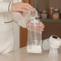 Someone pours breast milk from a small container into Nuby Breast Milk Storage Bags on a kitchen counter, with a breast pump nearby.