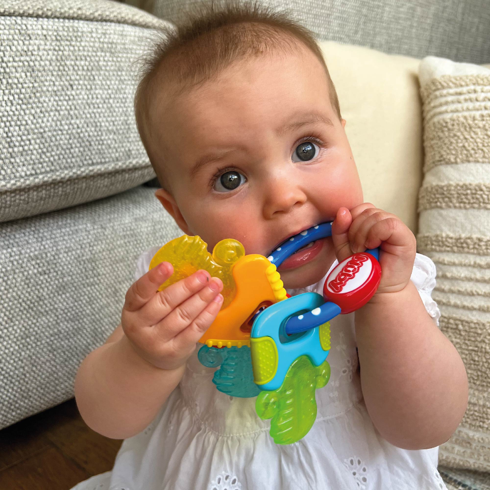 A baby in a white outfit sits on the floor, holding and chewing on the Nuby Icy Bite Keys Teether Toy, with a beige couch and striped pillows in the background.