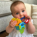 A baby in a white outfit sits on the floor, holding and chewing on the Nuby Icy Bite Keys Teether Toy, with a beige couch and striped pillows in the background.