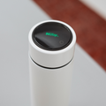 A close-up of the Nuby UK RapidCool Bottle Maker and Portable UV Steriliser Bundle, a white cylindrical device with a black circular digital display featuring the green Nuby logo. The blurred background shows gray and reddish-brown tones.
