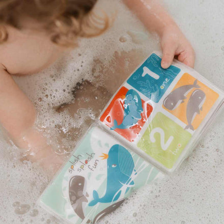 A young child enjoys a bubble bath while holding the Bathtime Book, featuring colourful whale illustrations and numbers one and two to support sensory development and early learning. These particular pages are coloured blue, green, yellow and red while floating in the bath itself.