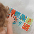 A young child in a bathtub holds the Bathtime Book, featuring colorful sea creatures and numbers five and six, promoting sensory development and playful learning.