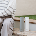 A person in a striped sweater and cargo pants sits on a bench next to two Nuby UK RapidCool Twin Pack and Portable UV Steriliser Bundle bottles, with green grass visible in the background.