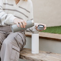 A person sits on a wooden bench outside, pouring water from a gray bottle into the Nuby UK RapidCool Twin Pack and Portable UV Steriliser Bundle.