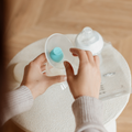 A person assembles the Nuby Silicone Manual Breast Pump with a blue silicone valve over a small white table, surrounded by a baby bottle, plastic bags, and a milk collector.