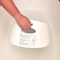 A hand holds the Newborn Baby Bath Support—made of white plastic—over a bathtub filled with water, displaying the instructions, labels, and maximum water level indicator on the back of the support.